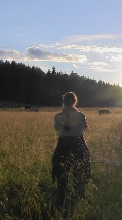 A woman in traditional clothing at a meadow and a cow grazing in the distance.